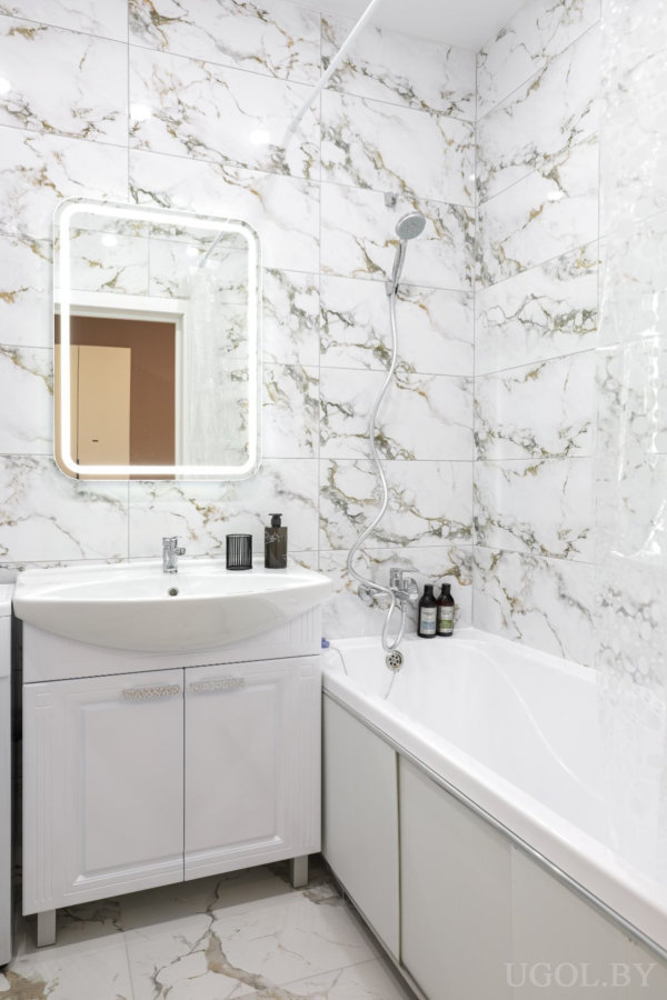 Современная ванная с мраморными стенами и полом, белой тумбой с раковиной, подсвеченным зеркалом и ванной с душем и косметикой на бортике.