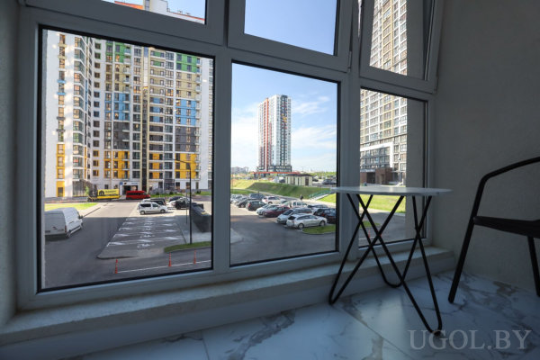 Панорамный вид через большие окна на яркие многоэтажные дома, парковку, зелёные зоны и ясное небо, внутри стол и стул на мраморном полу.