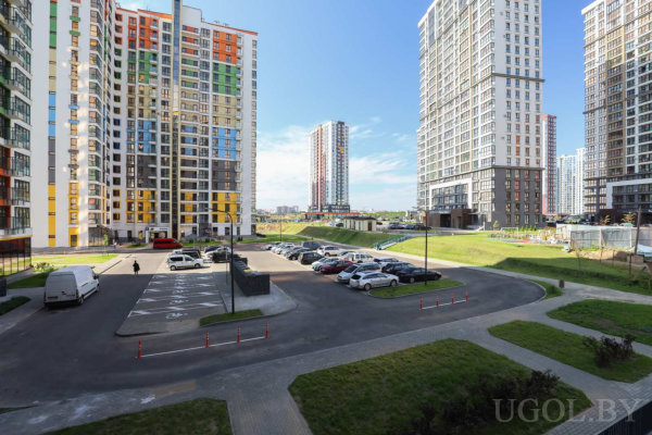 Высотные дома с цветными фасадами, парковка с машинами, пешеходы, зелёные газоны и уличное освещение в солнечную погоду.