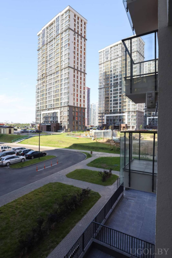 Панорамный вид с балкона с стеклянным ограждением на современные многоэтажные дома, парковку, зелёные зоны и пешеходные дорожки.