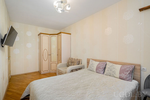 Уютная спальня с двуспальной кроватью, креслом, двухцветным шкафом и настенным телевизором.