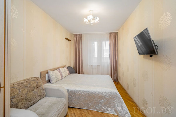 Минималистичная спальня с бело-серебристым покрывалом, телевизором на стене и светлыми шторами.