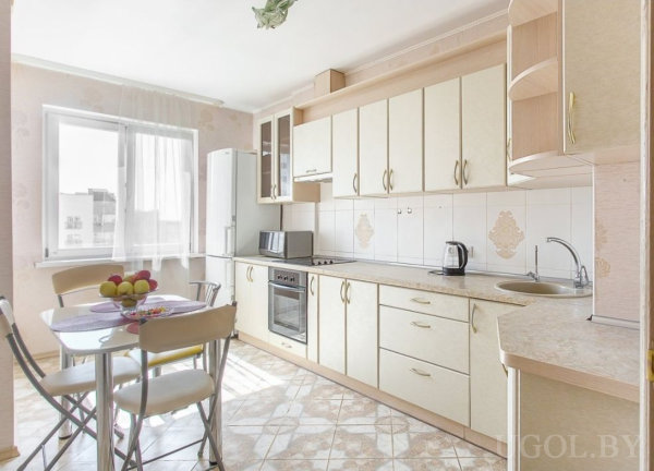 Светлая кухня с плитой, духовкой, холодильником и столом на четыре персоны у окна.