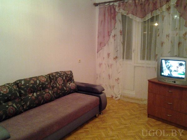 Небольшая гостиная с фиолетовым диваном с цветочным узором, деревянной тумбой с телевизором и окнами с розовыми шторами.