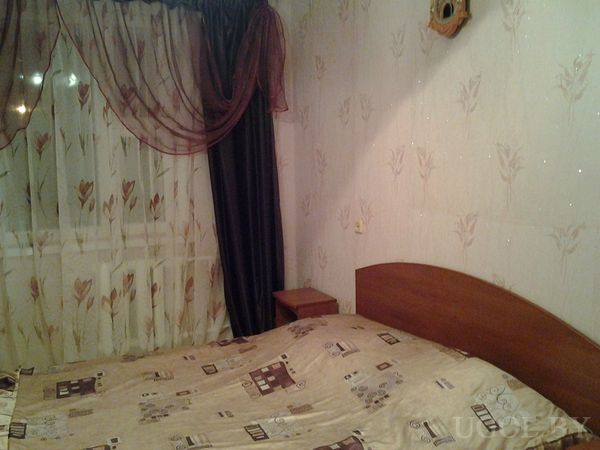 Уютная спальня с деревянной кроватью, покрывалом с геометрическим узором, прикроватной тумбой и настенными часами.