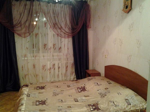 Комната с деревянной кроватью, бежевым покрывалом с узором, белыми и бордовыми шторами и настенными часами.