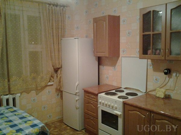 Небольшая кухня с белым холодильником, плитой, деревянными шкафами и столом с клетчатой скатертью.