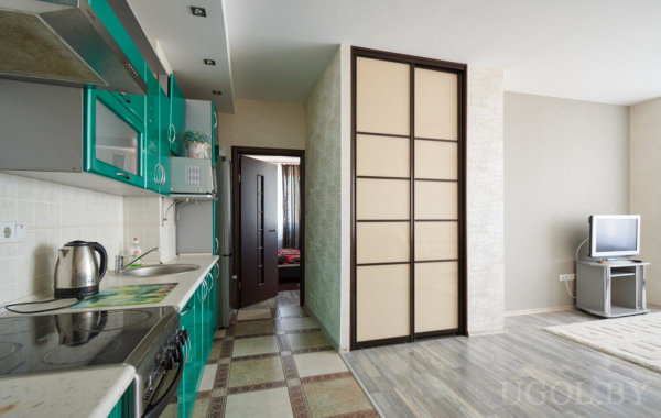 Интерьер с зоной кухни в бирюзовых тонах, телевизором на тумбе и проходом в спальню с красным покрывалом.