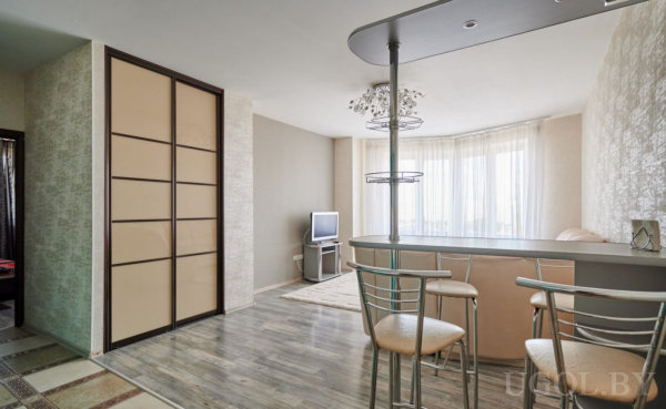 Светлая комната с телевизором, шкафом-купе и барной стойкой с металлическими стульями под подвесной декор.