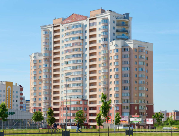 Здание с балконами и надписью «КВАРТИРЫ», окруженное зеленью и людьми, на фоне ясного неба.