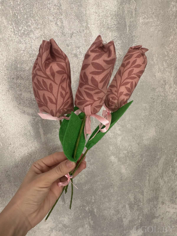 Рука держит три розовых цветка из ткани с зелеными листьями, перевязанные лентой на фоне серой стены.