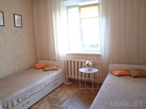 Две кровати с бежевыми покрывалами и оранжевыми подушками, столик с цветами и окно с роллетой.