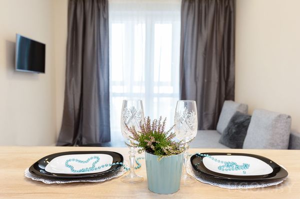 Обеденная зона с тарелками, бокалами, растением, серыми шторами и диваном на фоне.