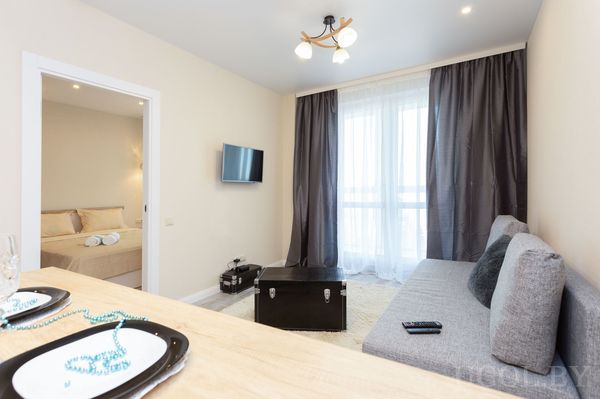 Интерьер с диваном, столом, телевизором, сервировкой, шторами и проходом в спальню.