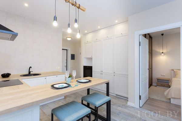 Композиция с белыми фасадами, деревянной столешницей, сервировкой, подвесными лампами и кроватью в глубине.