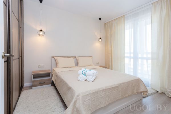 Комната с кроватью, тумбами, полотенцами, шторами, ковриком и шкафом с деревянными дверцами.