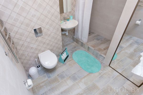 Интерьер с унитазом, умывальником, душем, корзиной, ковриком и светлой плиткой.