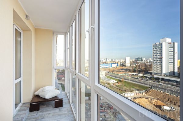 Застеклённая зона с лавкой, подушками, плиткой и видом на высотки и трассу.