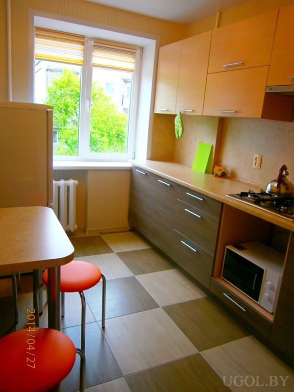 Компактная кухня с двухцветными фасадами, плитой, встроенной микроволновкой и обеденной зоной на фоне окна с жалюзи.