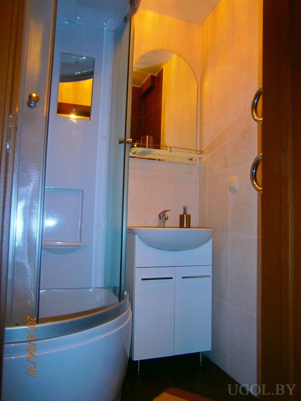 Угловая душевая с полками, белая тумба с зеркалом и мягкое освещение на фоне бежевой плитки.