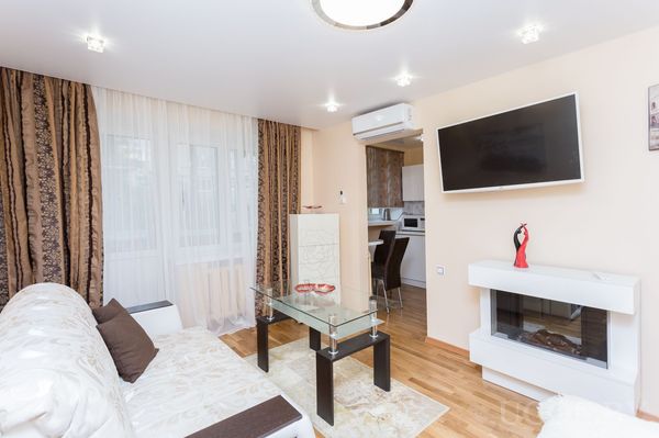 Современная гостиная с белым диваном, стеклянным столиком, телевизором над электрическим камином и коричневыми шторами.