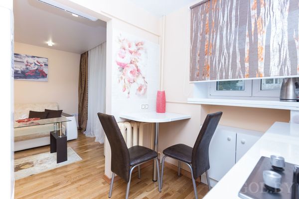 Компактная квартира с белой кухней, обеденным столиком у стены, красной вазой и гостиной с диваном.