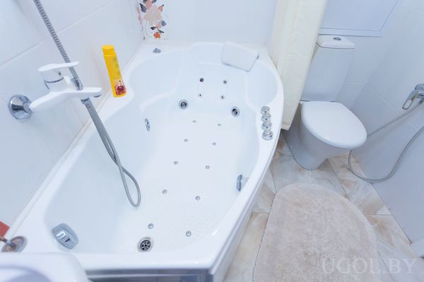 Ванная комната с угловой ванной‑джакузи, унитазом, бежевым ковриком и декоративной плиткой над ванной.