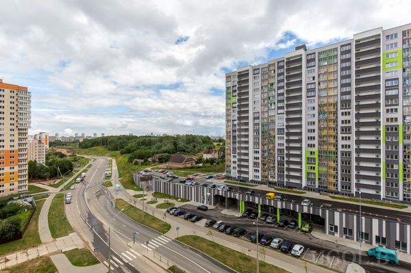 Уличная сцена с домами, парковкой, пешеходной зоной, деревьями и облачным небом.