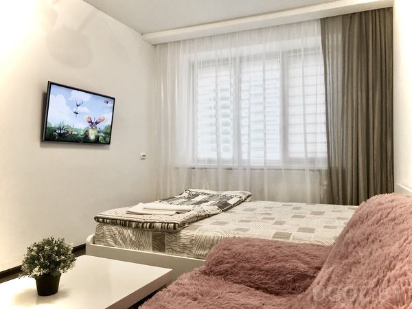 Современная спальня с кроватью, розовым креслом, белым столиком с растением и телевизором на стене.