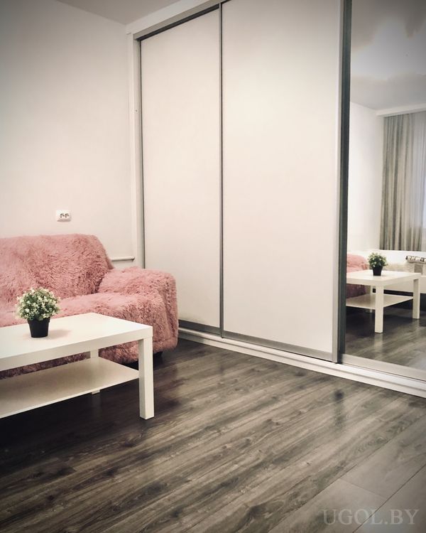 Светлая комната с розовым креслом, белым столиком с растением и большим шкафом с зеркальной дверцей.