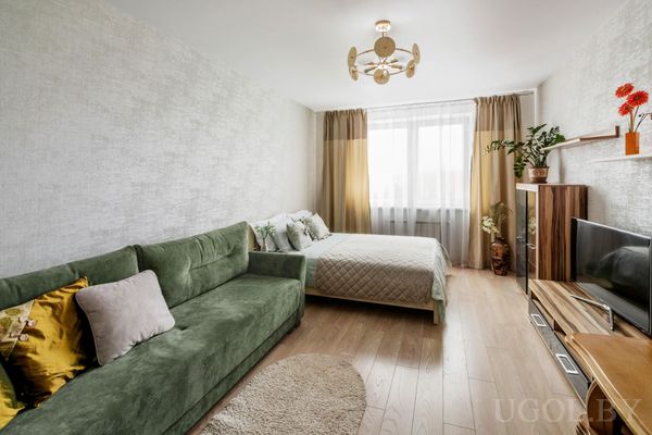 Жилая зона с зелёным диваном, кроватью с декоративными подушками, светлым ламинатом и тумбой с телевизором.