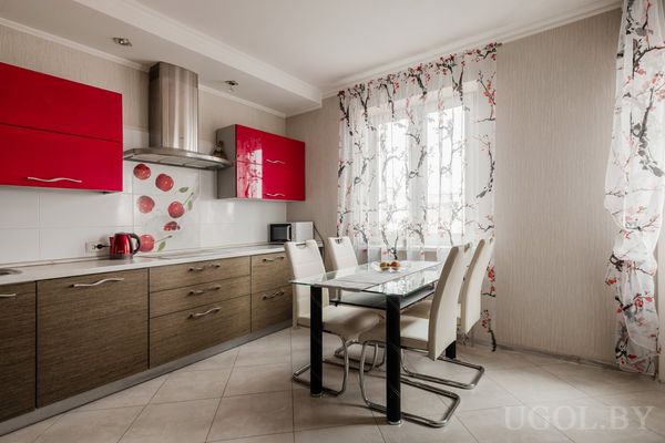 Кухонная зона с красными и деревянными шкафами, стеклянным столом и белыми стульями на фоне светлой плитки.