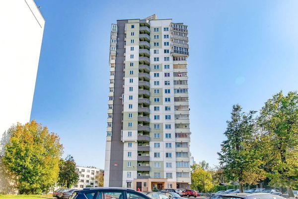 Жилой дом с бело-серым фасадом, балконами, деревьями и припаркованными машинами на фоне ясного неба.