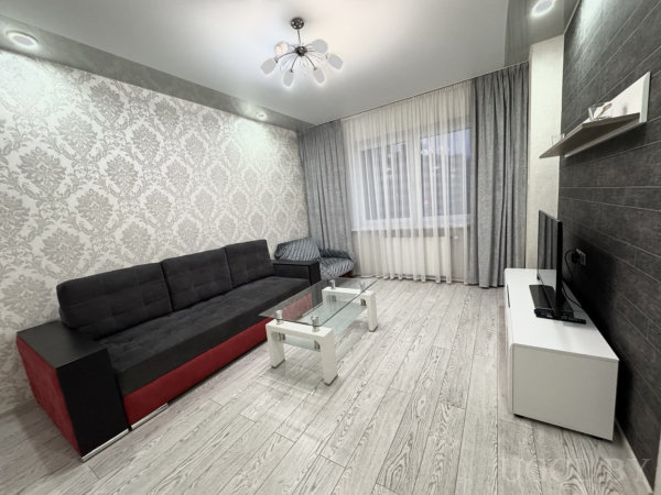 Комната с диваном, столом, телевизором, полкой, тюлем, серыми шторами и светильником на потолке.