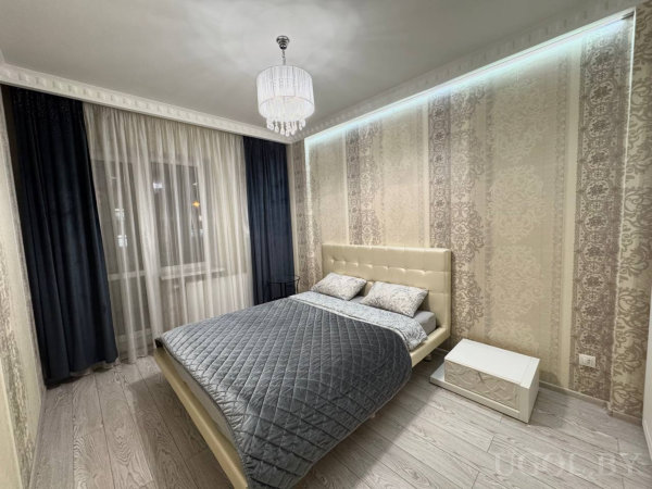 Интерьер с кроватью, бежевым изголовьем, тумбой, люстрой, тюлем, тёмными шторами и светодиодной лентой.