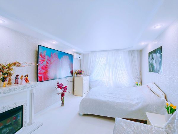 Комната с кроватью, белым камином, телевизором с изображением цветов, тюлем и декором в светлых тонах.