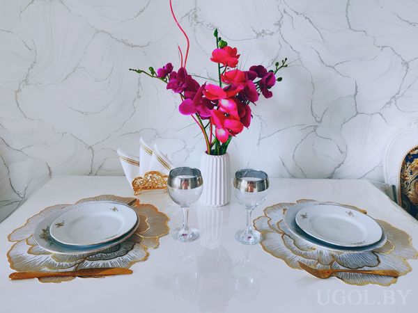 Стол с белыми тарелками, салфетками, серебряными бокалами и вазой с яркими цветами на фоне мрамора.