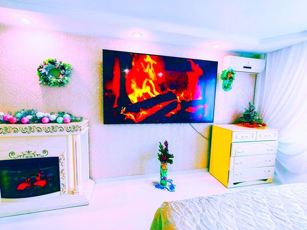 Интерьер с камином, телевизором с изображением огня, венками, ёлкой и белыми стенами.