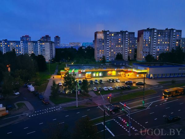 Уличная сцена с освещённым магазином, парковкой, автобусом, высотками и тёмно-синим небом.