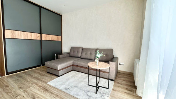 Секционный диван у светлой стены, круглый стол с вазой на ковре и шкаф-купе с тёмными панелями, лёгкие шторы у окна.