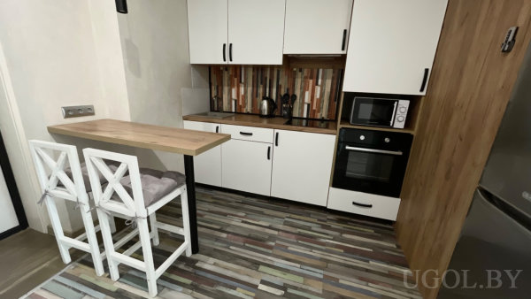 Белые шкафы с чёрными ручками, деревянная столешница, встроенные духовой шкаф и микроволновка в пенале, пёстрый вертикальный фартук и разноцветный деревянный пол.