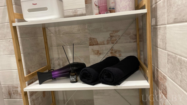 Деревянный стеллаж с белыми полками: фен, диффузор с палочками и чёрные полотенца, сверху туалетные принадлежности, контейнер «Dogi».