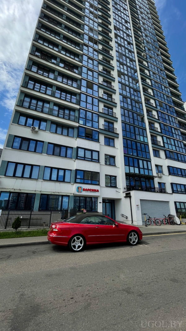 Высотка с белым фасадом и синим стеклом, у входа вывеска «BARCOWIA» с гербом, рядом припаркован кабриолет и велосипеды.