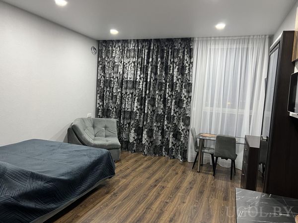 Современная студия с кроватью в синем текстиле, серым креслом, обеденной зоной у окна и тёмной тумбой с телевизором.