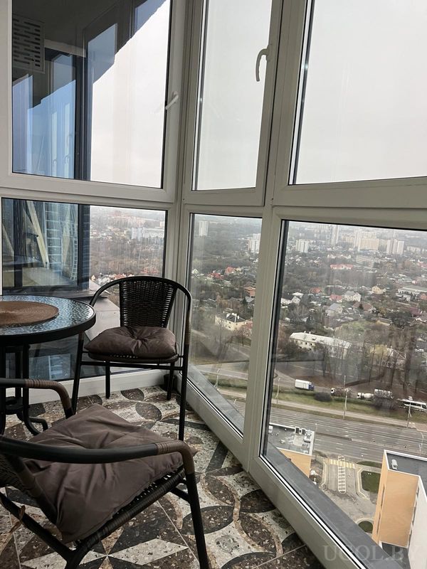 Застеклённый балкон с плетёной мебелью, стеклянным столиком и панорамным видом на городские дома и дороги.