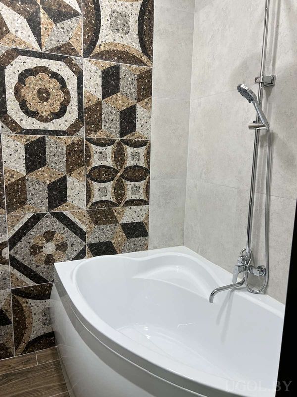 Угол ванной с белой ванной, душем на штанге и плиткой с геометричным узором на фоне серых стен.