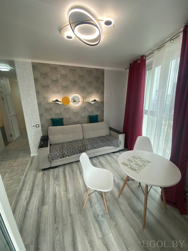 Комната с серым диваном, орбитальным потолочным светильником, декоративными полками и круглым столом у окна.