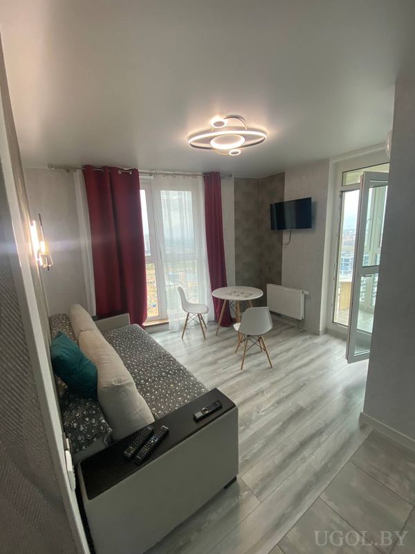 Комната с серым диваном, телевизором на стене, круглым столом и выходом на балкон с панорамным видом.