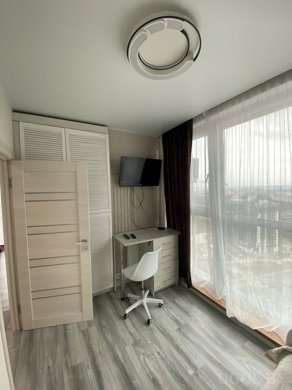 Комната с письменным столом, креслом, телевизором на стене и большими окнами с видом на высотки.