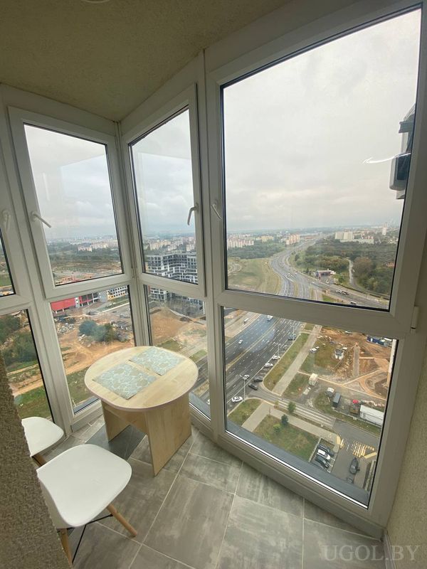 Застеклённый балкон с двумя белыми креслами, круглым столом и видом на дороги, стройку и высотки.